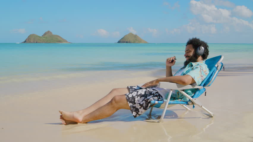 Black man lounges in a beach chair with headphones, smoking vape, enjoying a peaceful moment on a paradise tropical beach in Hawaiian island, relaxed and content. Oahu, Hawaii, Slow Motion, 4K RAW.  Royalty-Free Stock Footage #3442523117