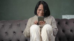 Elderly woman asian sit on couch in living room with cell phone, wear glasses looks at smartphone screen, web surfing information enjoy easy modern tech usage spend free time at home. 