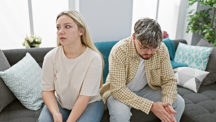 A worried young woman and a despondent man sit separately on a sofa in a modern living room, reflecting relationship troubles. Royalty-Free Stock Footage #3442690981
