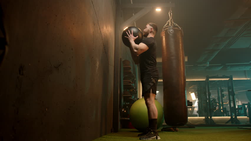 In the dimly lit expanse of an industrial gym, a man exerts full-body force in a medicine ball slam, the echo amplifying his intense focus on building muscular and cardiovascular power. 