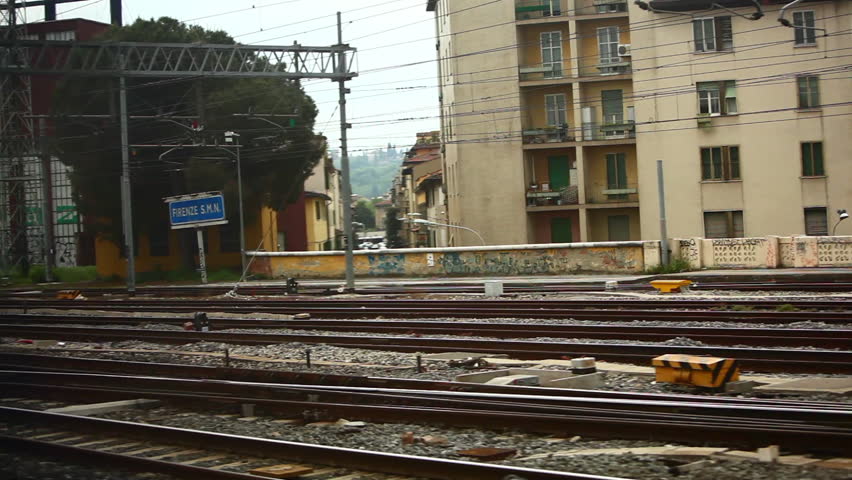A tracking shot of railway tracks in the Firenze S.M.N. station in Italy. Railway workers and a passing locomotive are seen and there are houses in the background. 