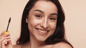 Close-up of a cheerful young woman applying mascara, showcasing a daily beauty routine with a neutral beige studio background. - Powered by Shutterstock - Get 15% off with code: PIKWIZARD15