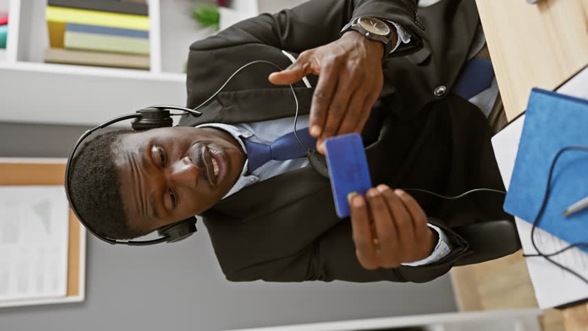 An african man in a suit with headphones on smiling at a blue card in a modern office.
