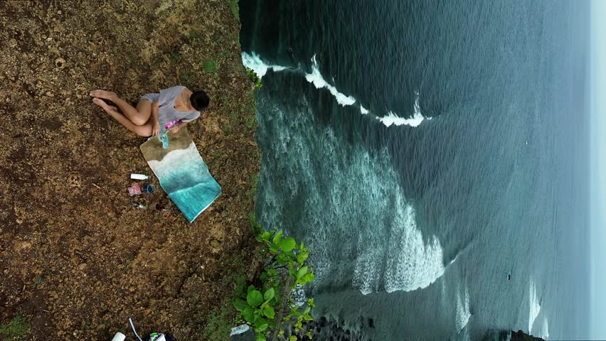 Aerial view of a woman painter who sits on the edge of a cliff near the ocean and draw a picture on canvas. She paints a seascape. She holds a palette in her hands and mixes blue paints with a brush.