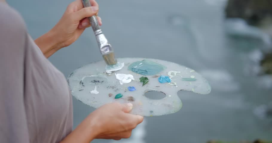 A close-up of a woman hands mixing different shades of blue acrylic paints with a brush on a palette. The cliff and ocean are visible in the background. The artist paints a picture near the sea.