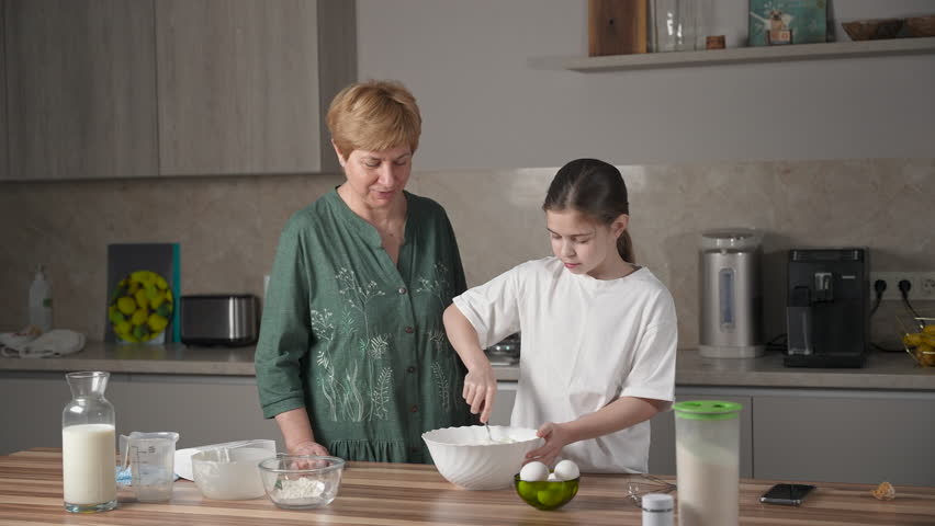 Lovely grandmother teach smiling granddaughter to make dough for pancakes for breakfast. Kissing child, having fun and enjoying time together on family kitchen at home on women's day Royalty-Free Stock Footage #3442806039