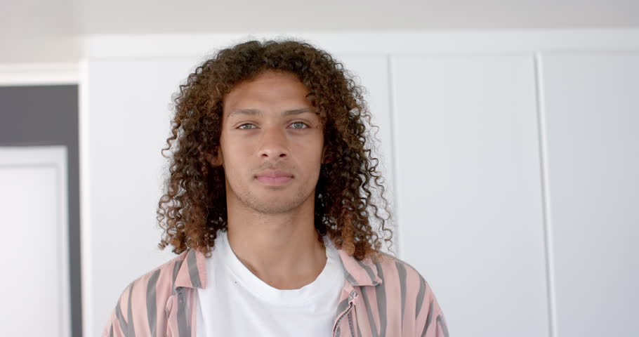 Young biracial man smiles confidently at home. His casual style and warm setting suggest a relaxed and welcoming atmosphere, slow motion.