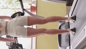 Vertical video: A young girl with long blond hair turns on the treadmill and begins to walk along it. Back view.