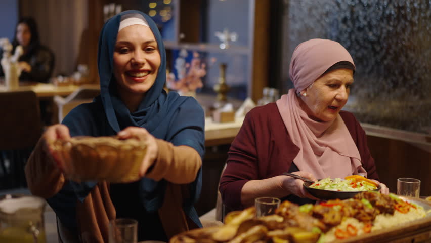 Muslim Woman in Hijab Sitting Next to Her Mother Giving Flatbread to Other Family Members at the Table in a Restaurant During Ramadan Iftar Meal or Eid Dinner with Meat in front of Her Royalty-Free Stock Footage #3442979103