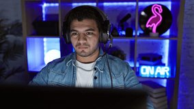A distressed man with headphones in a gaming room looks troubled by the computer screen glow during the night.