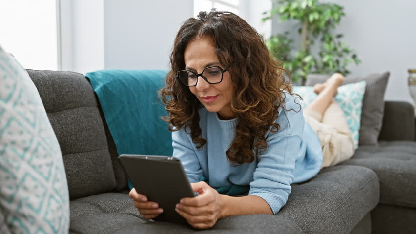 A mature hispanic woman with curly hair and glasses comfortably browsing on a tablet while seated on a grey sofa in a cozy living room. Royalty-Free Stock Footage #3443046009