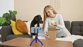 A young blonde woman at home plays with her black dog, recording a video indoors with a smartphone on a tripod.