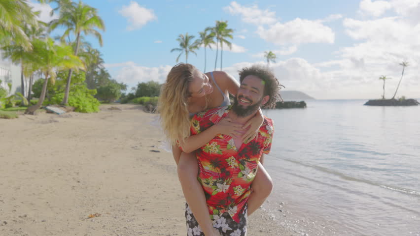 African American man carries a caucasian woman on his back, enjoying Hawaiian beach with a picturesque landscape with palm trees and clear blue sky at the island of Oahu, Hawaii, USA Royalty-Free Stock Footage #3443293131