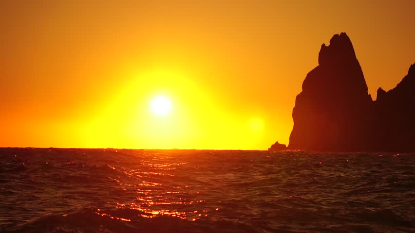 Red burning sunset over stormy sea with rocky volcanic cliff. Abstract nature ocean sea background. Small waves on golden warm water surface with bokeh lights from sun. Weather and climate change