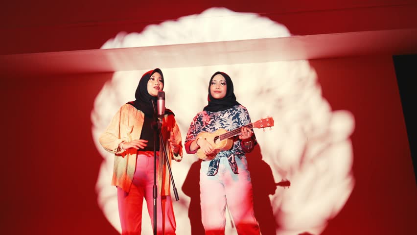 Two people on stage, one holding a microphone and the other a guitar, with a focus on cultural diversity during the night