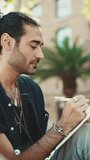 VERTICAL VIDEO, Young italian guy with ponytail and stubble sits on street bench and makes sketches with pen on piece of paper
