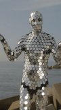 A man in a full body disco suit dancing next the sea in vertical video format