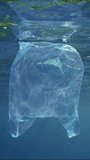 Vertical video, Close-up of disposable blue plastic bag floating in blue water in bright sunshine, slow motion. Plastic pollution of Ocean, Discarded transparent blue plastic bag drifts underwater