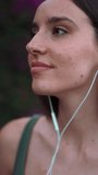 VERTICAL VIDEO: Clouse-up, woman with freckles and dark loose hair wearing green top walks along wall blooming with purple flowers. Pretty girl listening to music with wired headphones