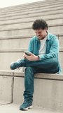 VERTICAL VIDEO: Smiling young bearded man in denim shirt sitting on high steps and using mobile phone. Man looking at his smartphone social networks. Slow motion