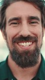 VERTICAL VIDEO, Close-up portrait of a smiling man with a beard on the embankment. Frontal closeup portrait of happy young hipster male looking at camera