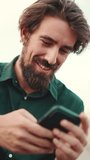 VERTICAL VIDEO, Close-up of smiling man with a beard chatting on the embankment background. Frontal closeup of happy young male hipster using mobile phone