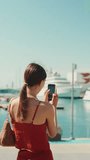 VERTICAL VIDEO: Close-up cute girl takes photos, videos of the seaport on a mobile phone. Girl uses social networks and streaming services on a smartphone when shooting yachts and ships