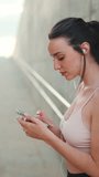 VERTICAL VIDEO: Young athletic woman with long ponytail wearing beige sports top in wired headphones, stands with mobile phone in her hands