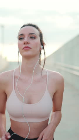 VERTICAL VIDEO: Young athletic woman with long ponytail wearing beige sports top in wired headphones, runs along the pedestrian crossing to the beach Royalty-Free Stock Footage #3443914645