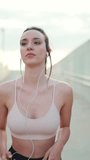 VERTICAL VIDEO: Young athletic woman with long ponytail wearing beige sports top in wired headphones, runs along the pedestrian crossing to the beach
