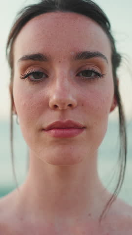VERTICAL VIDEO: Close-up young athletic woman with long ponytail wearing beige sports top is standing eyes closed and relaxing on the sea background