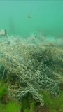 Vertical video, Lost fishing net lies on seabed in green algae Ulva in sun glare on shallow water, Slow motion. Ghost gear, fishing gear that has been abandoned, lost or otherwise discarded