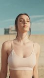 VERTICAL VIDEO: Young athletic woman with long ponytail wearing beige sports top walks along the beach at dawn on modern city background