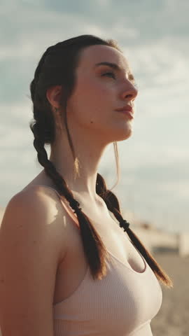 VERTICAL VIDEO: Young athletic woman with braided pigtails wearing beige sports top walks along the beach at dawn and looks at the sea Royalty-Free Stock Footage #3443915597