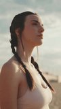 VERTICAL VIDEO: Young athletic woman with braided pigtails wearing beige sports top walks along the beach at dawn and looks at the sea