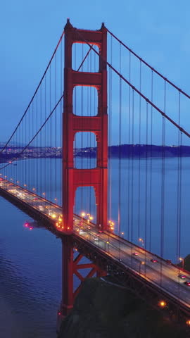 Vertical video. San Francisco landmark at sunset on cloudy day, aerial view of the Golden Gate Bridge and city skyline. Cinematic drone footage of red bridge with night illumination on blue sky