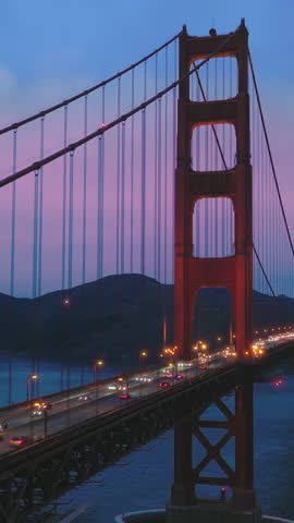 Vertical video. Aerial view of illuminated Golden Gate Bridge at pink sunset. Cloudy sunset above night traffic on red bridge over blue Pacific Ocean waters. San Francisco Bay, California, USA.