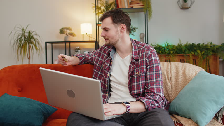 Young sad man guy in plaid shirt sitting on sofa using laptop and credit card frowns trying to make payment through internet feels angry due lack of money bankruptcy. Caucasian victim of fraud concept Royalty-Free Stock Footage #3443932947