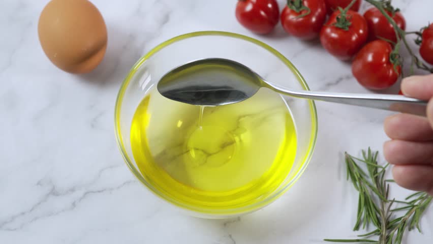A hand stirs with a spoon in a bowl of golden olive oil, on a table with fresh vegetables and an egg Royalty-Free Stock Footage #3443999431