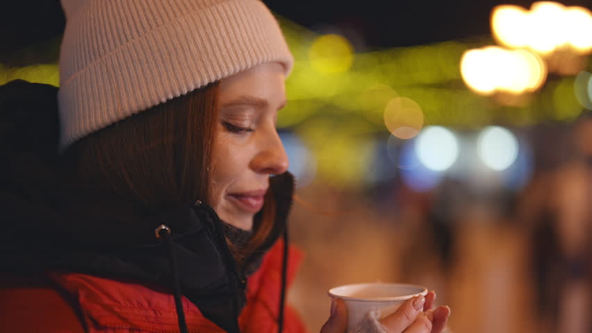 Closeup face of happy pretty young woman drinking hot wine at Christmas market in Europe. Cute female wears white hat drinking hot coffee or chocolate on blurred background bright bokeh festive lights