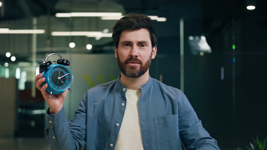 Portrait of handsome bearded man directing finger on vintage alarm clock and smiling at camera. Confident office worker reminding about importance of conscious time management for productivity.