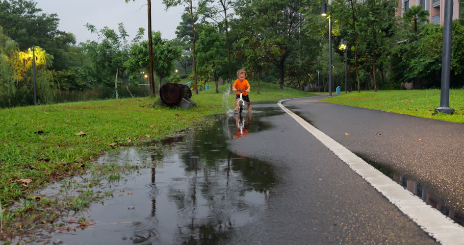 Joyful toddler races on a balance bike through puddles in a park after rain, creating splashes of water. Carefree escapade captures innocence and dynamic energy of childhood play in drizzling weather