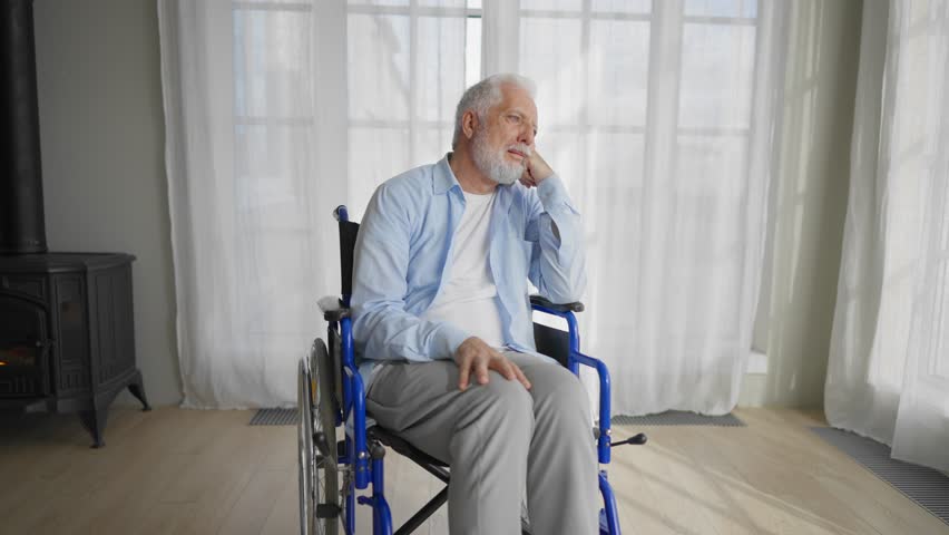 Old man in wheelchair looks at window. Thoughtful gray haired senior male at home enjoying pastime leisure. Living with physical legs trauma paralysis, sadness, loneliness of older people concept.