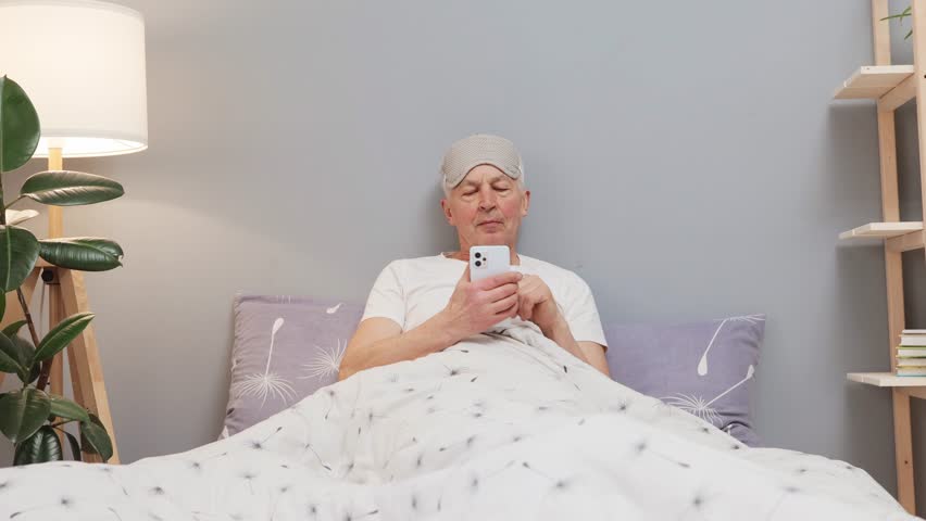 Awake and browsing. Seniors on social media. Mobile apps for the elderly in retirement. Caucasian elderly man in sleeping mask lying on bed using smartphone at home