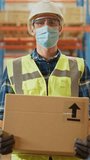 Handsome Male Worker Wearing Medical Face Mask and Hard Hat Carries Cardboard Box Stands in Retail Warehouse full of Shelves with Goods. Logistics, Distribution Center. Vertical Screen