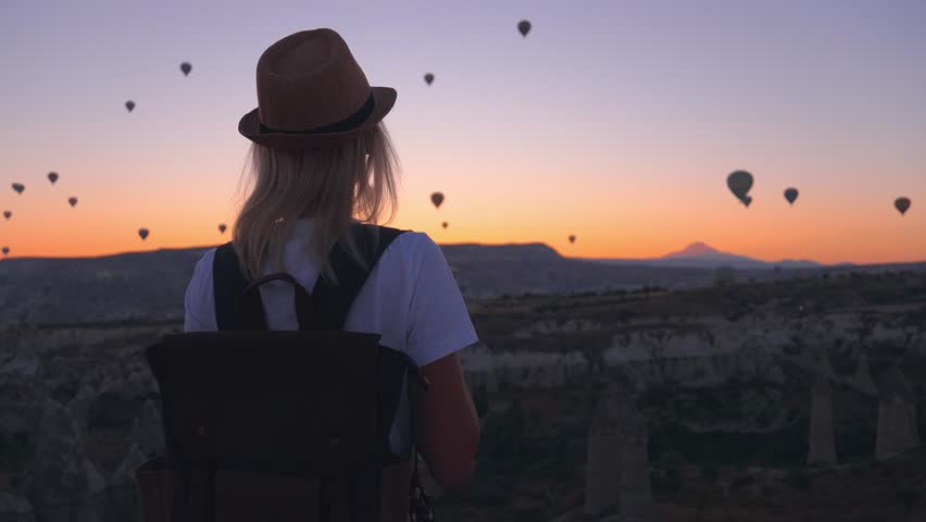 beautiful woman in hat backpack walking towards fairytale view balloons at sunrise in Gorem Cappadocia Turkey. Image of traveler and wanderer, she looks at fabulous and beautiful sunrise in mountains