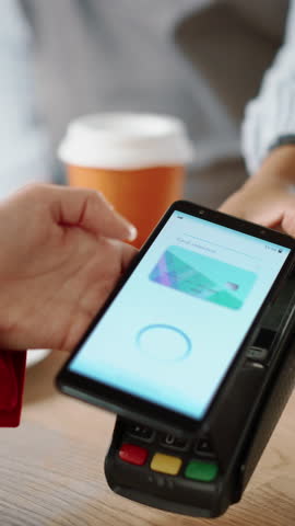 Vertical Footage of a Close Up of a Hand Holding a Bank Terminal Used for NFC Payment Technology on Smartphone. Customer Uses Mobile to Pay for Take Away Latte to Barista in a Coffee Shop.