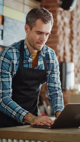 Vertical Footage of Handsome Coffee Shop Owner is Working on Laptop Computer and Checking Inventory in a Cozy Cafe. Restaurant Manager in Checkered Shirt Browsing Internet and Chatting with Friends. Royalty-Free Stock Footage #3444280247