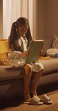 Vertical Screen: Portrait of a Little Korean Girl Sitting on a Couch in the Living Room and Using Tablet Computer to Play Video Games. Active Female Child Using Interactive Media for Learning