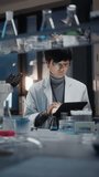Medical Development Laboratory: East Asian Scientist Uses Digital Tablet Computer, Looks Under Microscope, Conducting Experiment. Pharmaceutical Lab for Research Medicine, Biotechnology. Evening Work